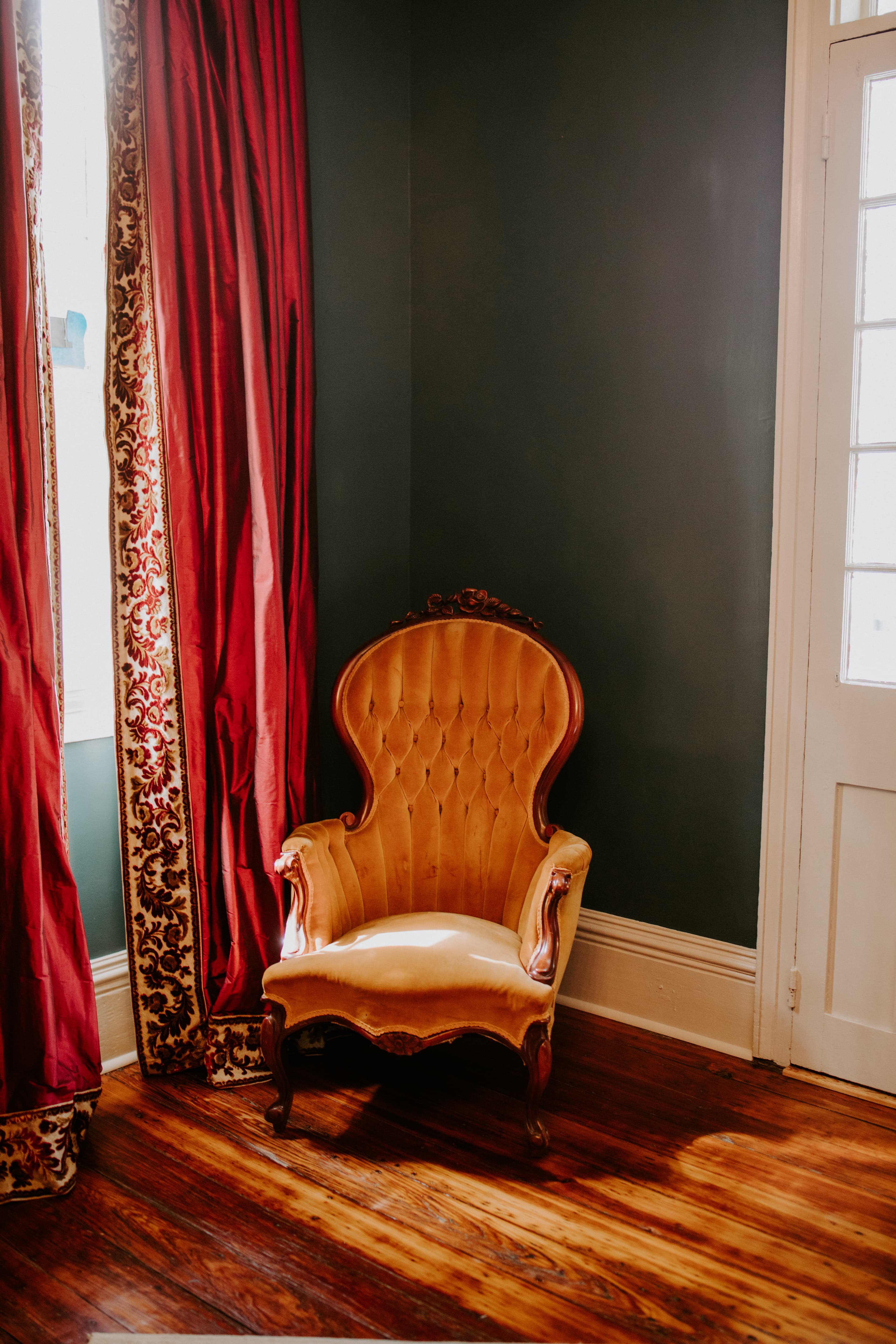 A vintage velvet armchair sits in a corner beside rich red curtains and a light-colored wall.