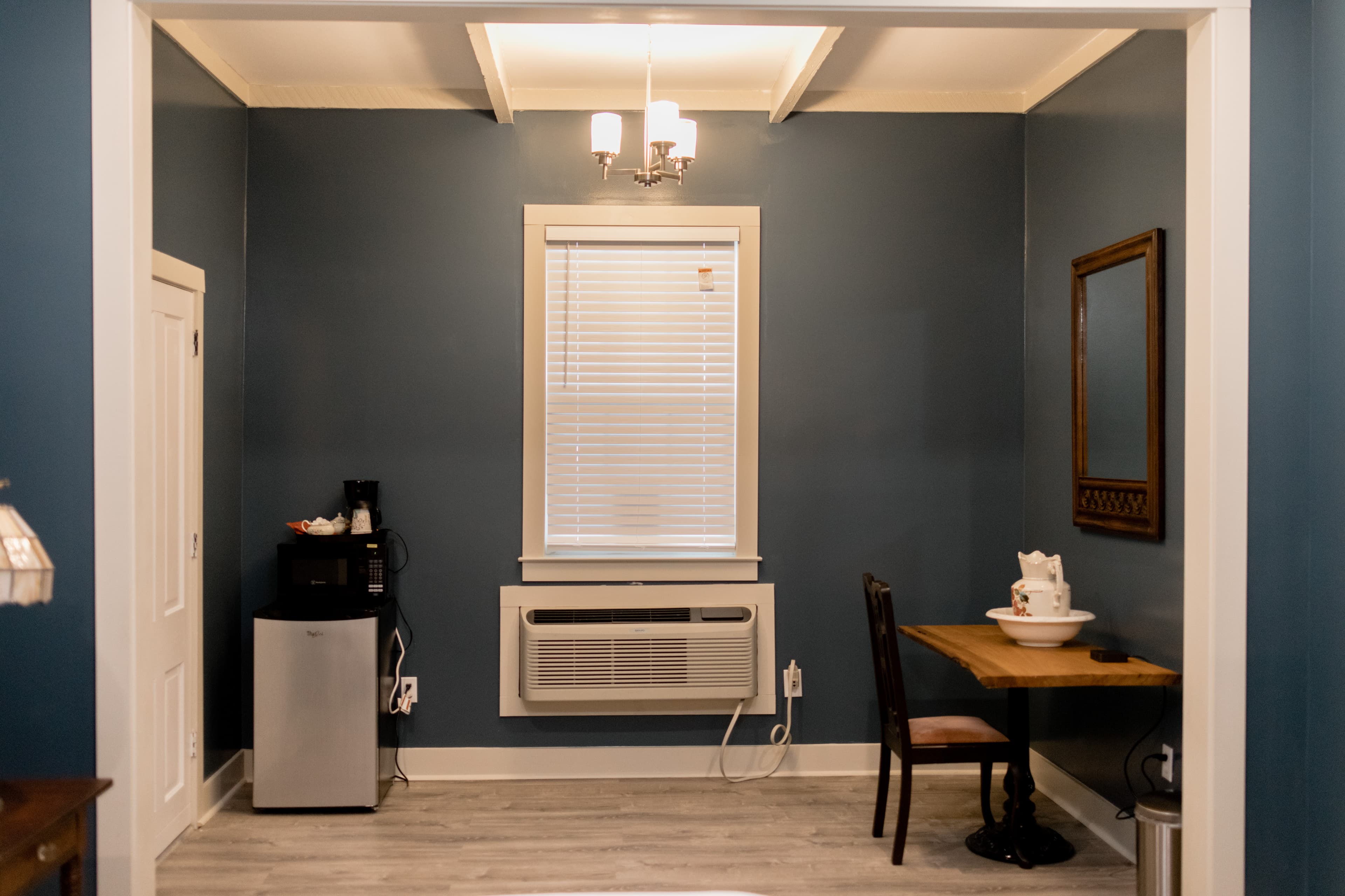 A cozy room featuring a small table with a chair, an air conditioning unit, a mirror, and a mini-fridge against a blue wall.