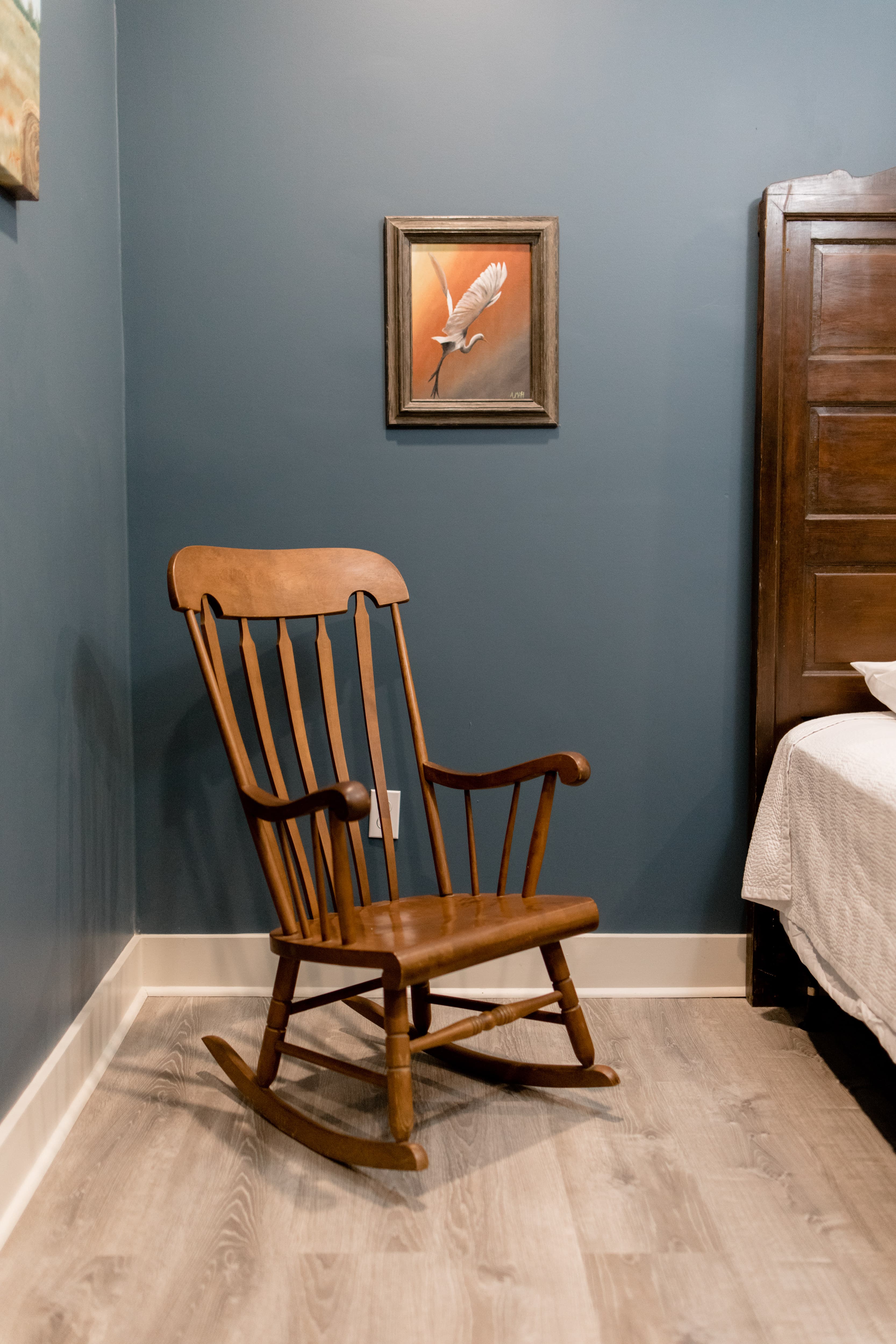 A wooden rocking chair stands against a dark blue wall, with a bird painting hung above it.