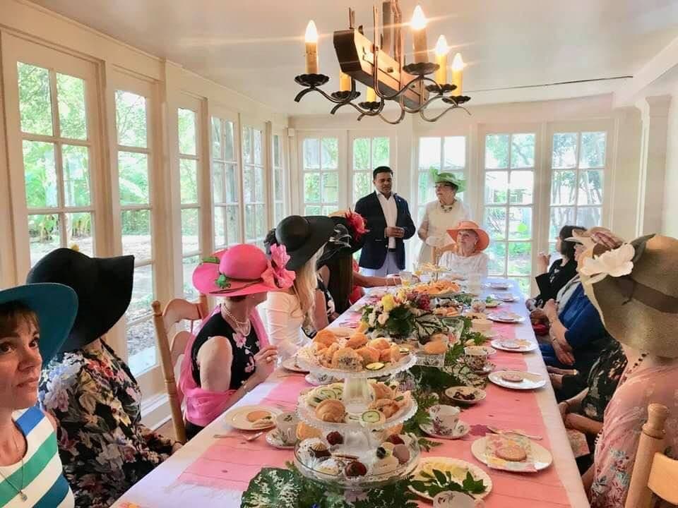 A group of women in stylish hats listens as a man and woman speak at a tea party set with an elaborate spread of food on a long table.