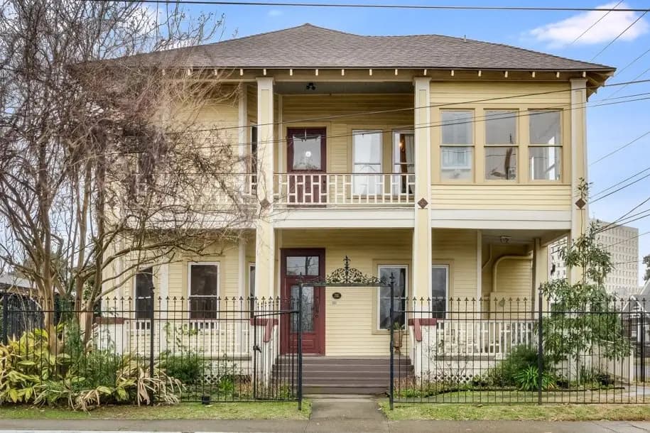 A two-story yellow house with a front porch, surrounded by a black fence and greenery.