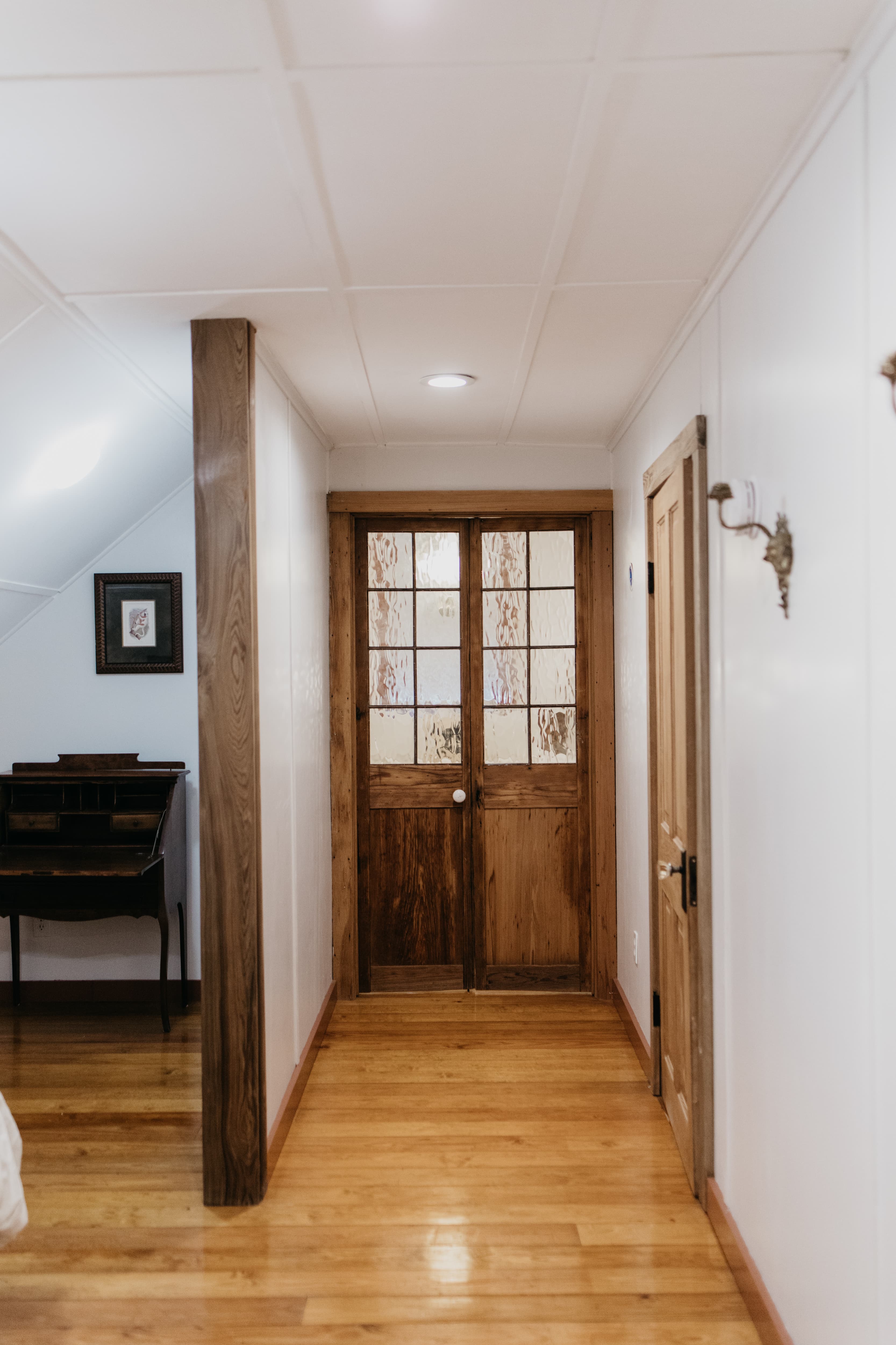 A hallway featuring a wooden double door at the end, lined with white walls and wooden flooring.