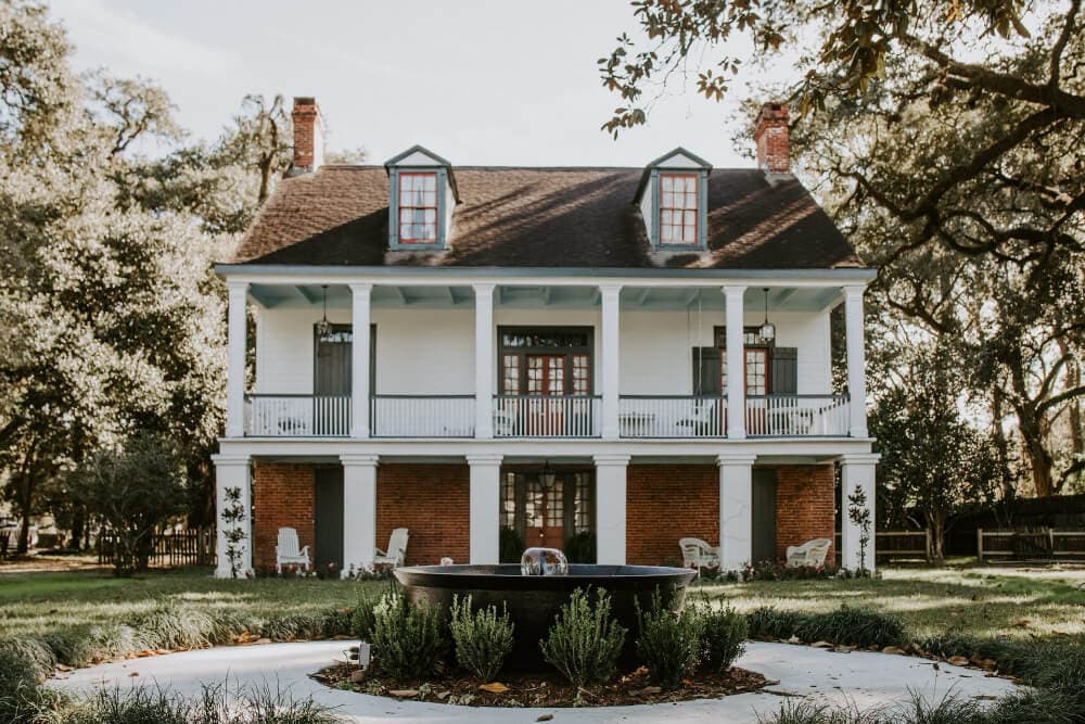 A historic two-story house with a porch, surrounded by greenery and featuring a central fountain.