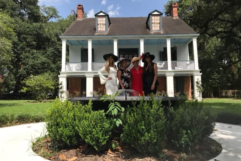 Four women in fashionable hats pose in front of a large historic house surrounded by greenery.