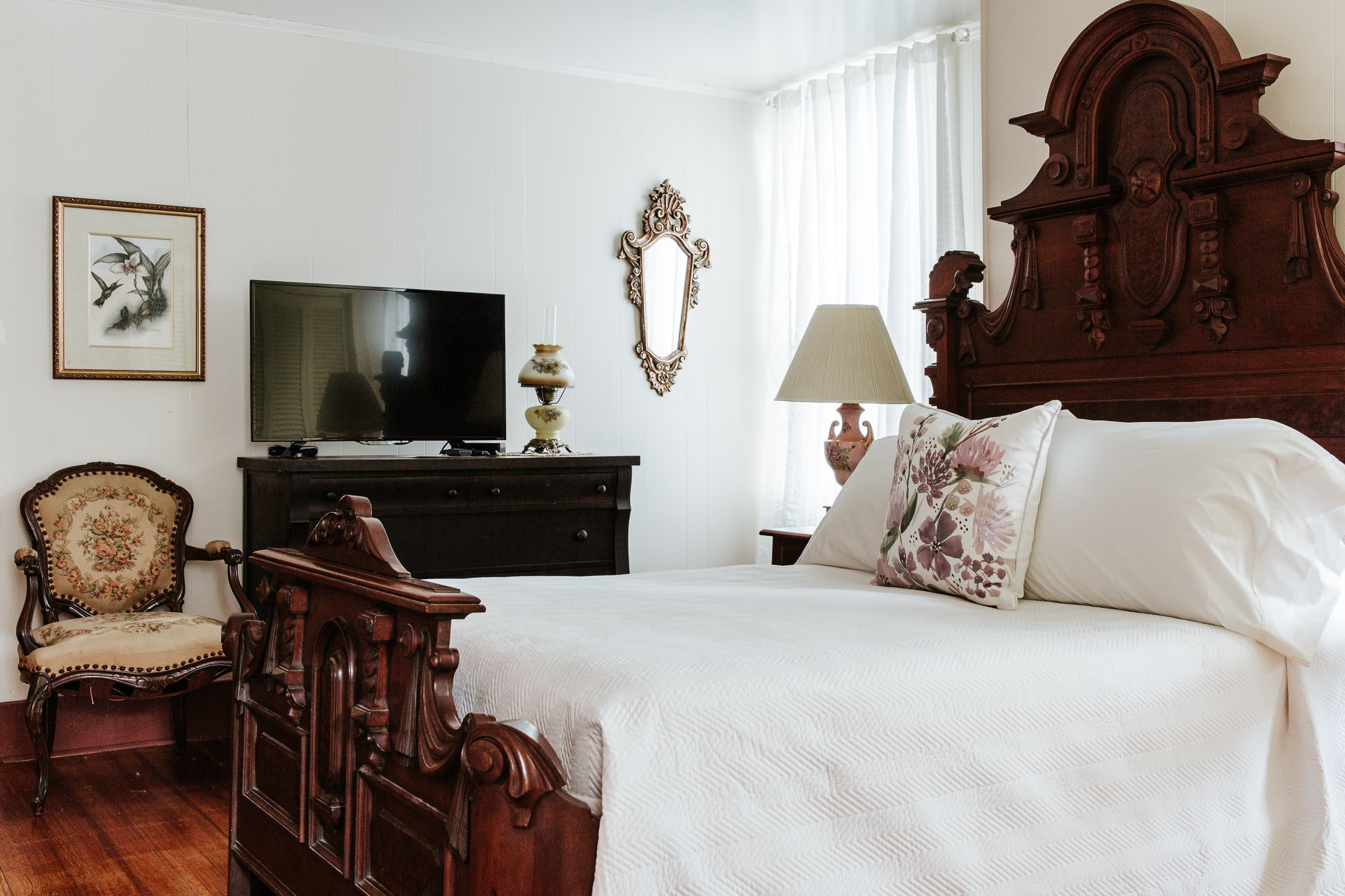 A cozy bedroom featuring a wooden bed, antique chair, television, and decorative mirror.