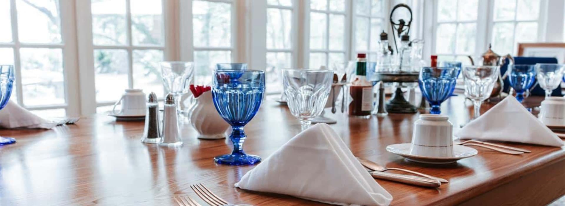 A dining table set with blue glassware and neatly folded napkins, surrounded by windows.
