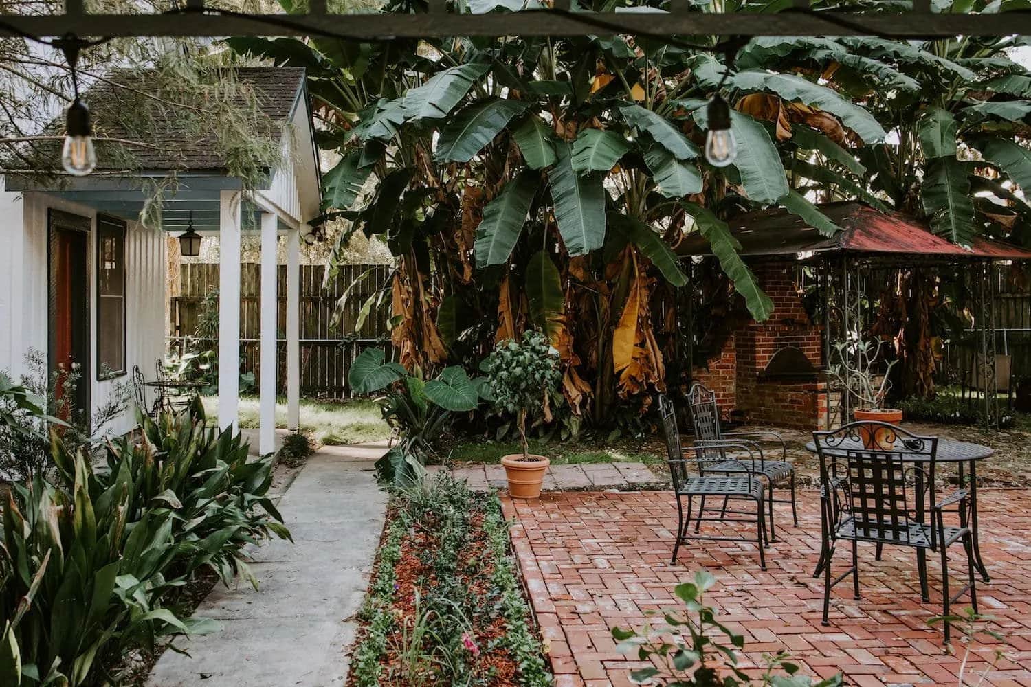A cozy garden patio features brick flooring, outdoor seating, and lush tropical foliage.